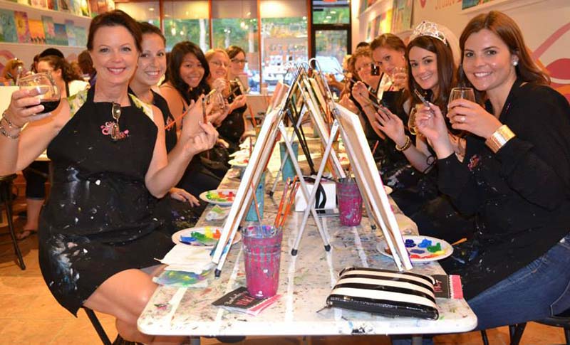 a group of women holding wine glasses & paint brushes at a paint & sip bachelorette party